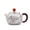 Sterling silver teapot with intricate floral relief, polished wood handle, and a round red stone finial.